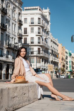 full length of brunette woman in skirt sitting on city street near handbag and straw hat clipart