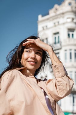 cheerful woman holding hand above eyes while looking away on street