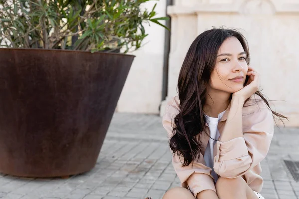 Esmer kadın çiçek saksısının yanındaki kaldırımda oturuyor ve başka tarafa bakıyor.