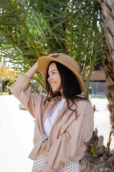 brunette woman in straw hat and beige shirt smiling under palm tree