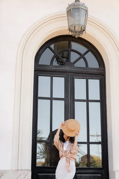cheerful woman obscuring face with straw hat near arch window