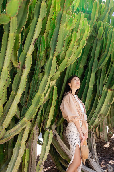 pleased woman in stylish clothes looking at giant succulents in park