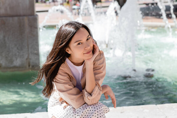 pleased woman looking at camera near fountain on blurred background