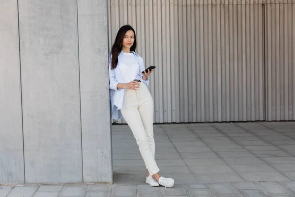 full length of brunette woman standing with smartphone and coffee to go near grey wall