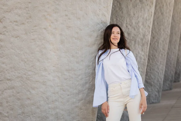 Smiling Brunette Woman Blue Shirt White Pants Leaning Grey Wall — 스톡 사진