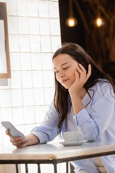 Dışarıdaki kafede kahve fincanının yanında cep telefonuna bakan pozitif bir kadın.