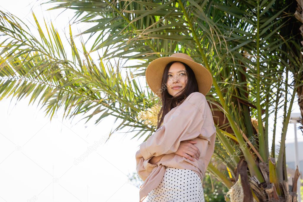 low angle view of happy brunette woman standing with crossed arms near palm tree