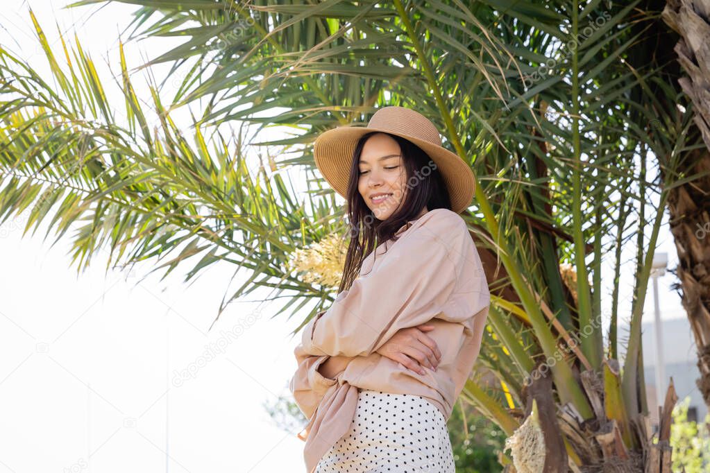 smiling brunette woman in straw hat standing with crossed arms under green palm tree