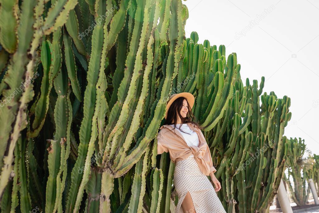 cheerful brunette woman in straw hat near green giant cactuses