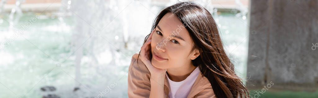 positive brunette woman holding hand near face while looking at camera outdoors, banner