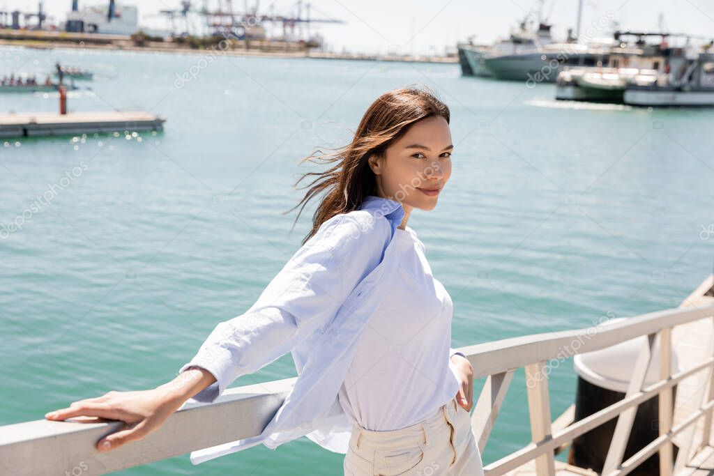 smiling woman in blue shirt looking at camera while standing on pier in seaport
