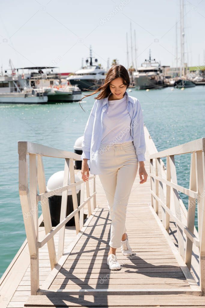 full length of smiling woman in blue shirt and white pants walking on pier in seaport