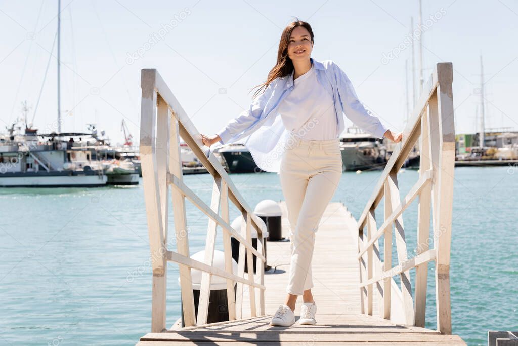 full length of smiling woman in white pants standing on pier in seaport