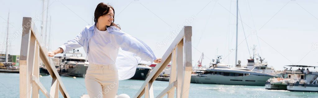 cheerful brunette woman looking away on pier in seaport, banner