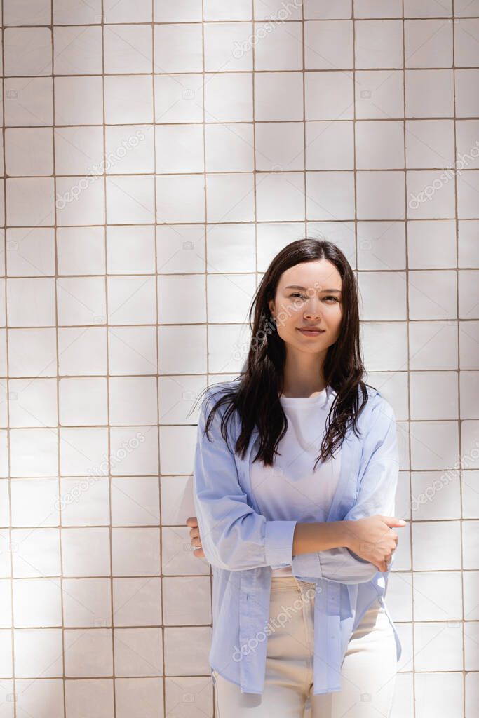 brunette woman in blue shirt standing with crossed arms near white tiled wall