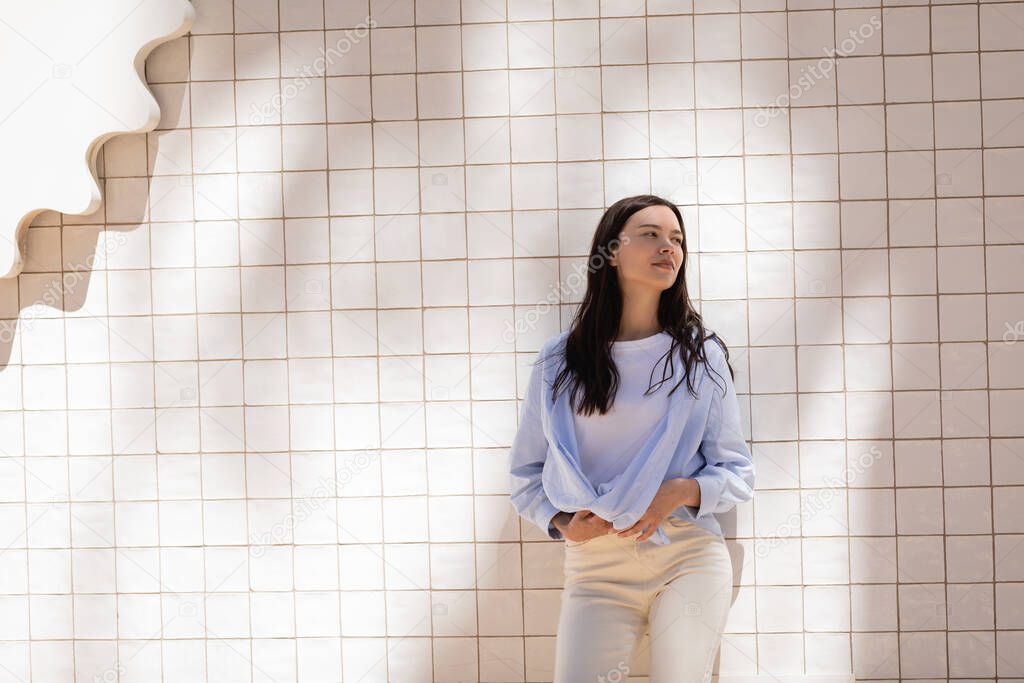 brunette woman in blue shirt standing near white tiled wall and looking away