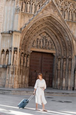 Valizli mutlu, kızıl saçlı bir kadın Valencia 'da bir binanın yanında duruyor.