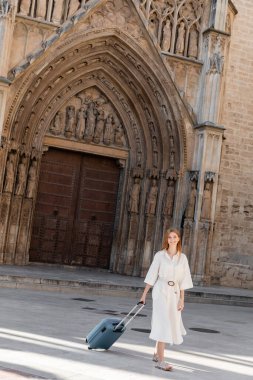 full length of cheerful redhead woman with luggage walking near building in Valencia clipart