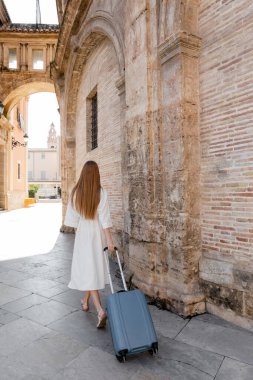 Beyaz elbiseli kızıl saçlı kadının arkadan görünüşü Valencia caddesinde bavuluyla yürüdüğünü gösteriyor.