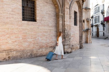 full length of redhead woman walking with luggage and using smartphone in valencia  clipart