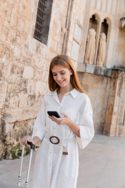 Valencia caddesinde elinde bavul tutmakta ve akıllı telefon kullanmakta olan neşeli kızıl saçlı kadın. 