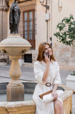 Valencia 'daki çeşmenin yanında beyaz elbiseli neşeli bir kadın akıllı telefondan konuşuyor.