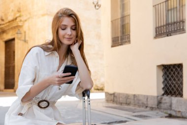 Beyaz elbiseli mutlu kızıl kadın bagaj koluna yaslanıyor ve Valencia 'da akıllı telefondan mesajlaşıyor.