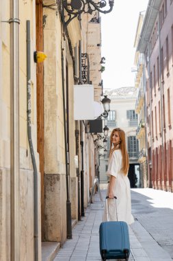 pleased redhead woman in dress standing with luggage on urban street in valencia clipart