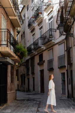 redhead woman in dress standing near houses on european street in valencia  clipart