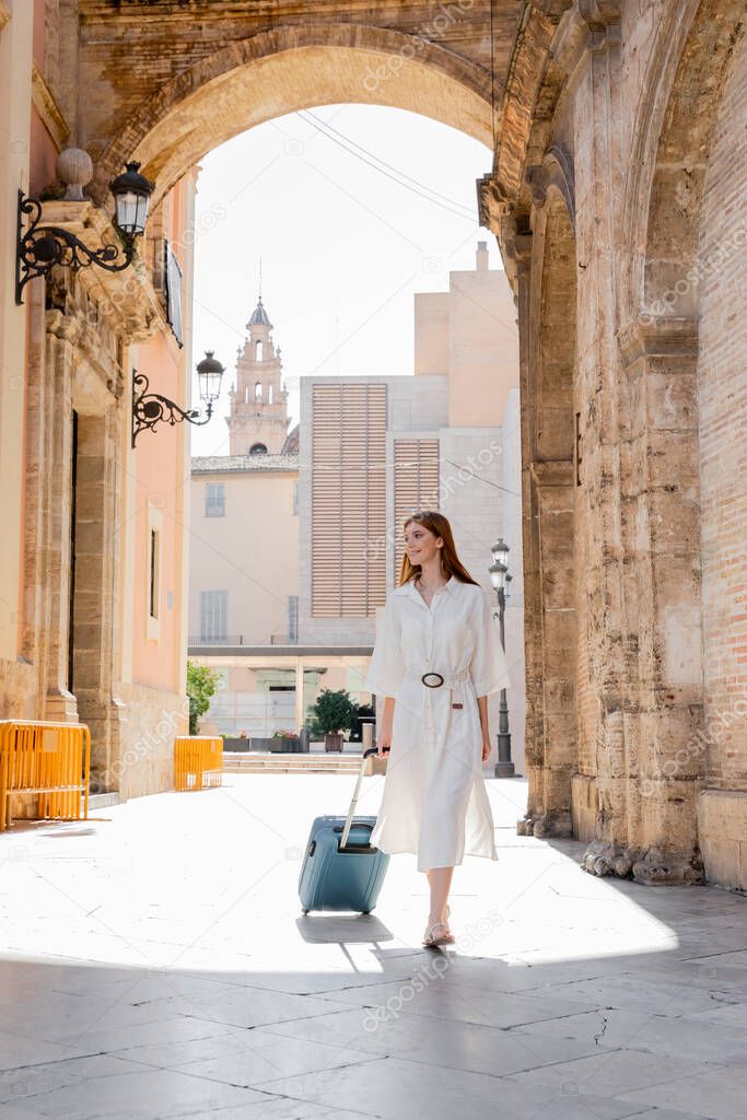 full length of cheerful redhead woman walking with baggage on street of Valencia