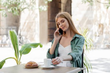 Kafe terasındaki masada ayçöreği ve kahve fincanının yanında akıllı telefondan konuşan kızıl saçlı kadın. 