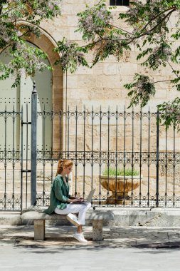 redhead woman sitting on concrete bench and using laptop near forged fence in valencia  clipart