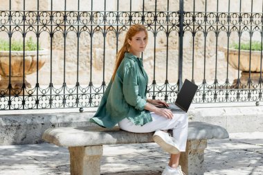 redhead freelancer sitting on concrete bench with laptop near forged fence in valencia  clipart