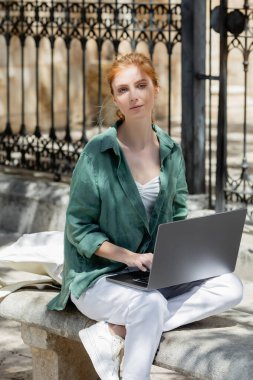 redhead freelancer sitting on concrete bench with laptop near forged fence  clipart