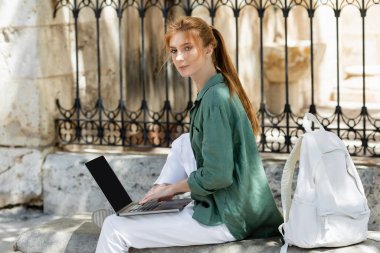redhead freelancer sitting on concrete bench and using laptop near backpack clipart