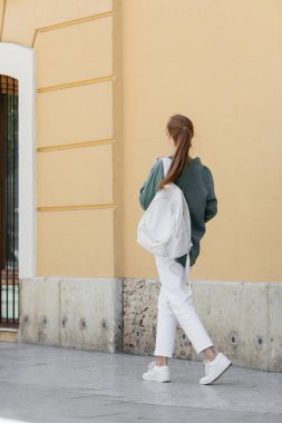 Yeşil tişörtlü, beyaz pantolonlu, sırt çantalı ve Valencia caddesinde bej duvarın yanında yürüyen kızıl saçlı bir kadın. 