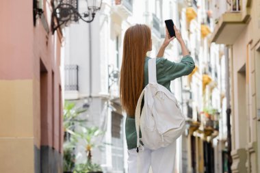 Sırt çantalı kızıl saçlı kadın Valencia şehir sokağında akıllı telefondan fotoğraf çekiyor.