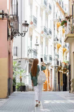 back view of redhead traveler with backpack taking photo on smartphone while walking on urban street of valencia clipart