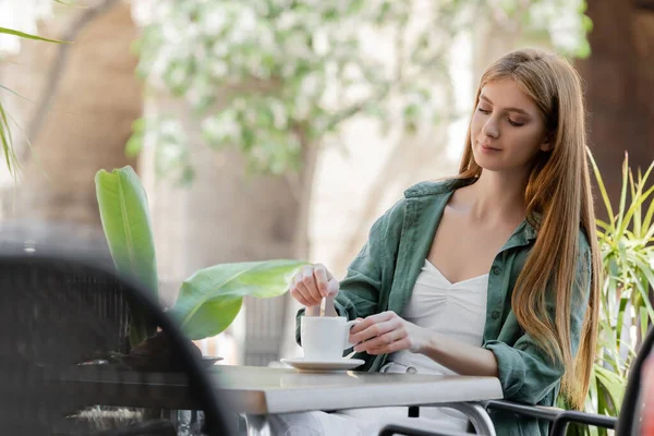 pleased woman stirring coffee near croissant on table in cafe terrace