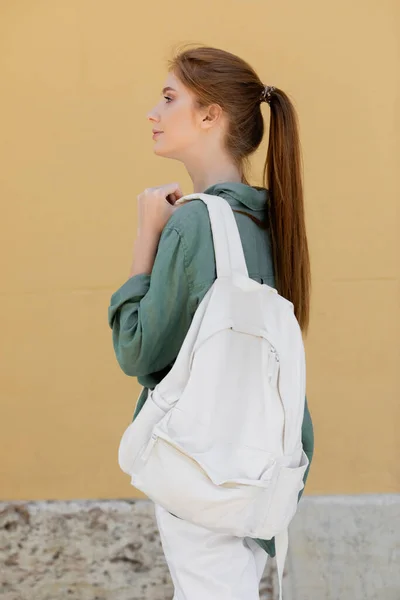 side view of young woman with red hair in green linen shirt holding backpack near beige wall on street of valencia