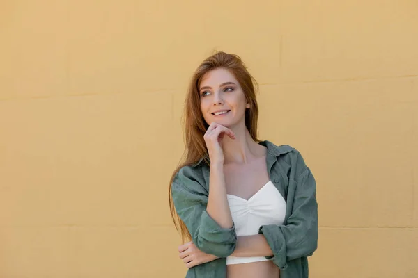 dreamy woman in green linen shirt smiling near beige wall on street