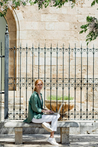 young redhead freelancer using laptop and sitting on concrete bench near forged fence in valencia 