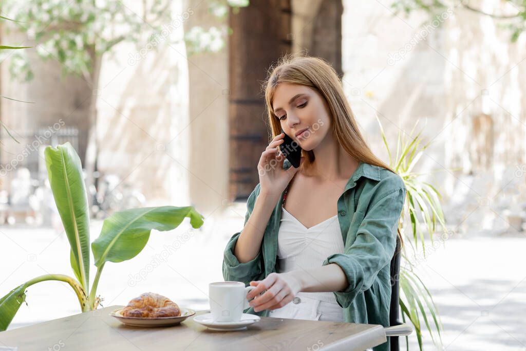 redhead woman talking on smartphone near cup of coffee and croissant on table in cafe terrace 