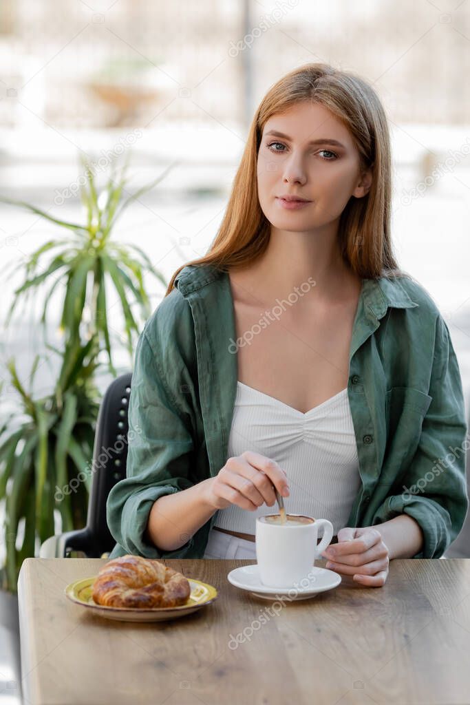 redhead woman stirring coffee with spoon and looking at camera on summer terrace