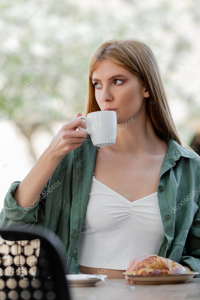 redhead woman drinking coffee near tasty croissant on summer terrace