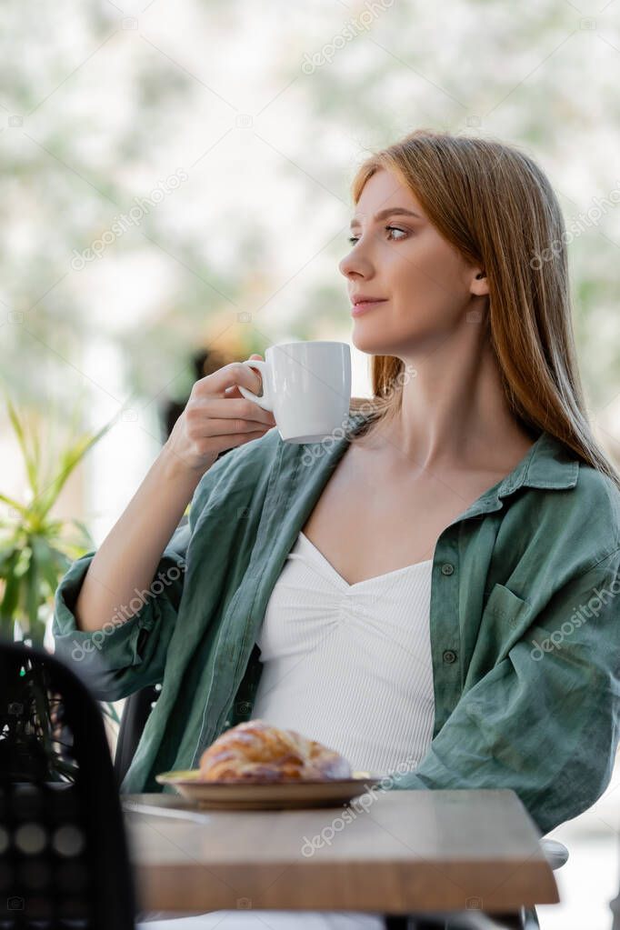 redhead woman holding cup of coffee near tasty croissant on summer terrace