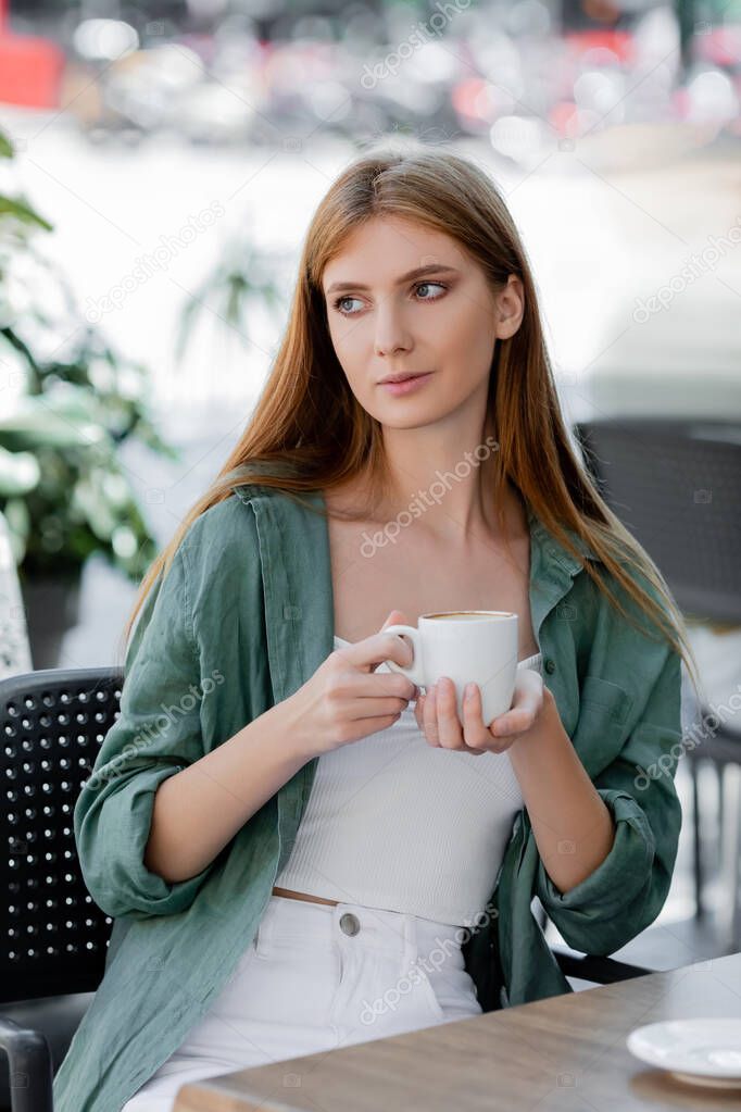 young woman with red hair holding cup while sitting in cafe terrace on european street