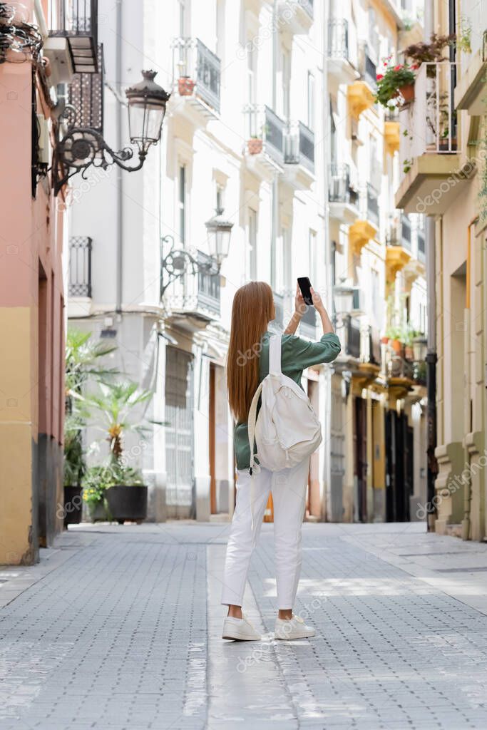 full length of redhead traveler with backpack taking photo on smartphone on urban street of valencia