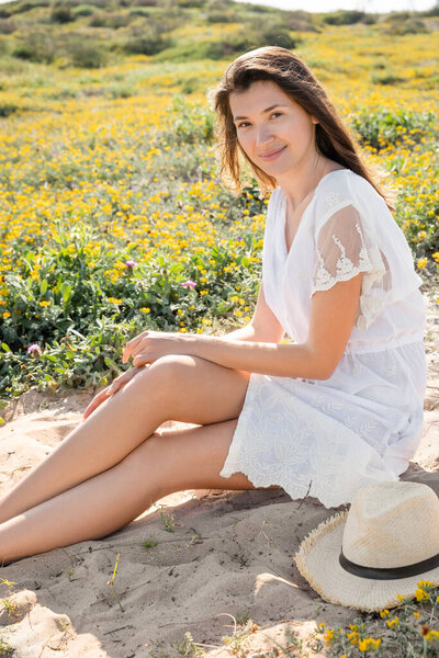 Positive woman in dress looking at camera near straw hat on beach near flowers 