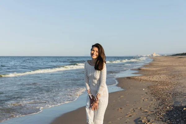 Positive brunette woman in knitted dress looking at camera near sea on beach